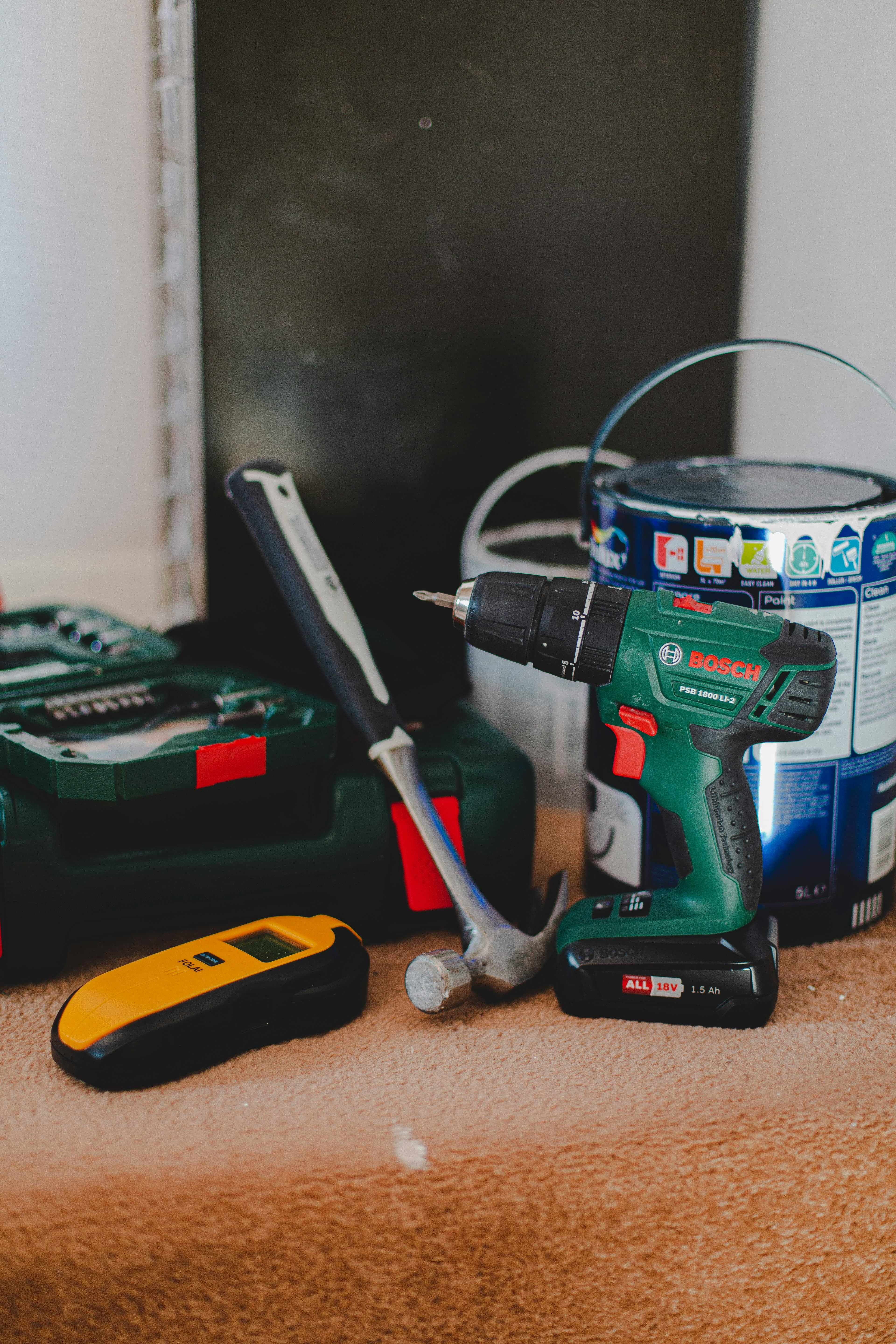 Green Bosch power drill, hammer, yellow stud finder, and paint can on brown carpet.